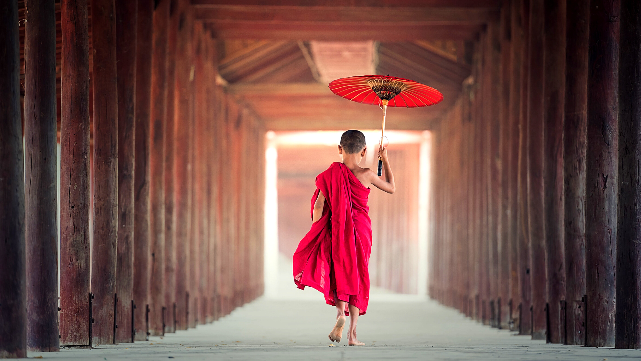 Young monk in red robes with a red umbrella, walking barefoot towards a brightly lit open door
